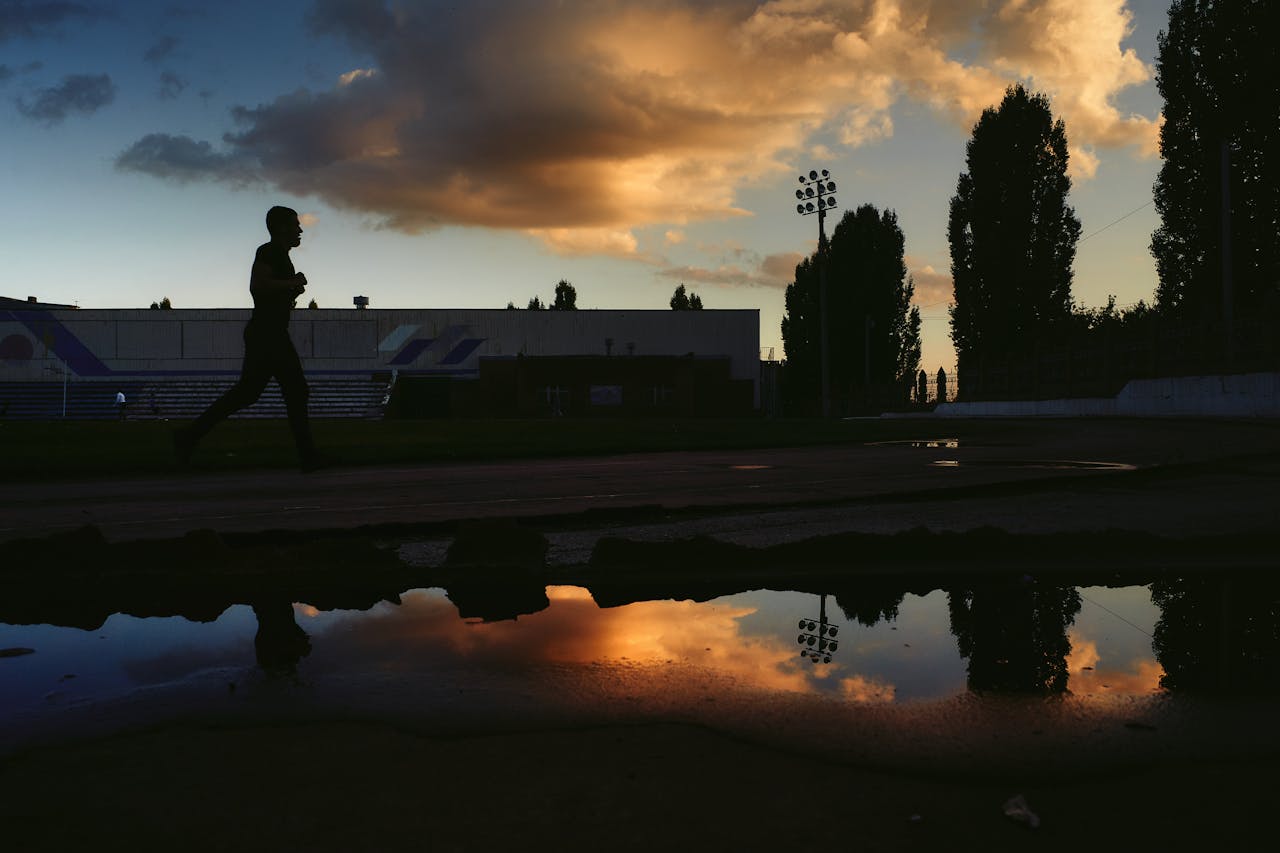 our-experience A silhouette of a man running on a track with a dramatic sunset sky reflected in puddles.