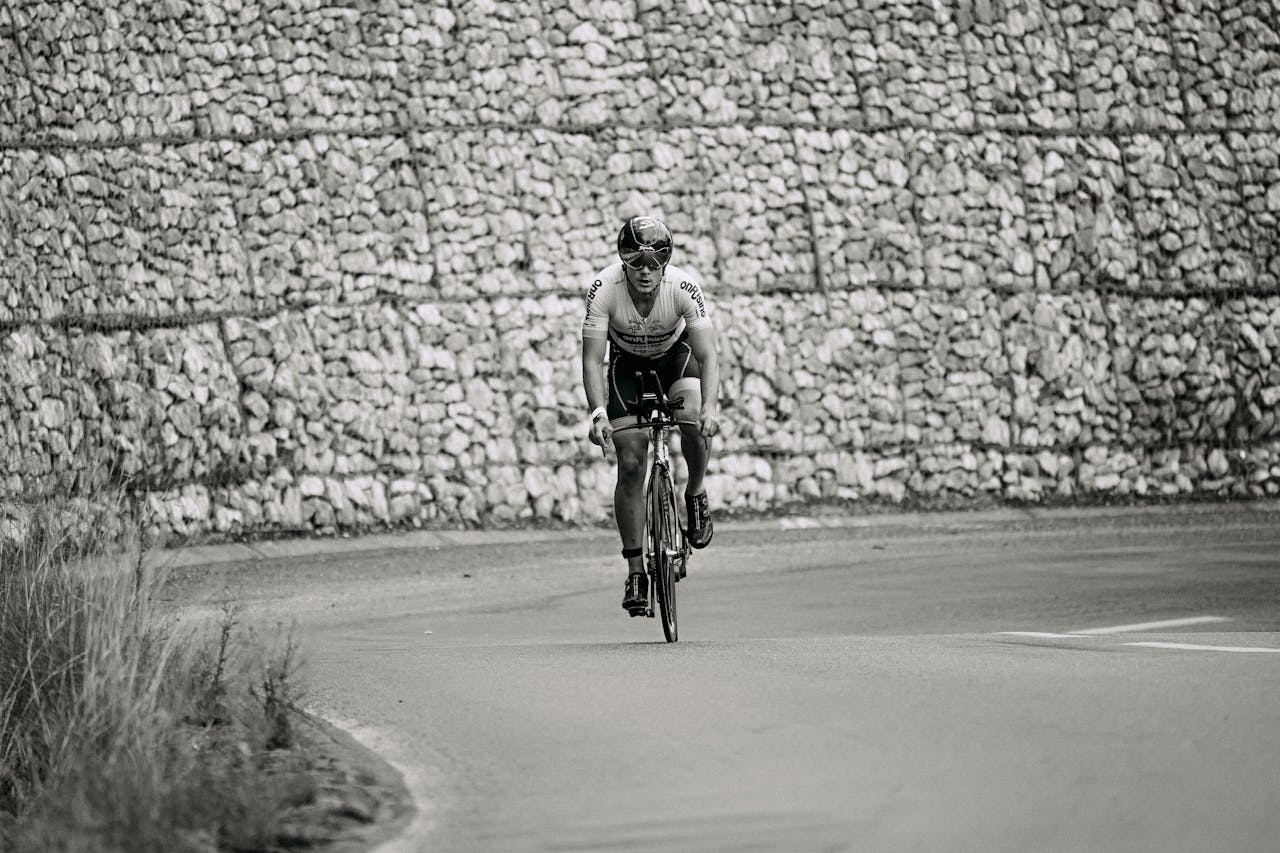 ours-journey A cyclist in sportswear riding along a curving road against a stone wall in grayscale.